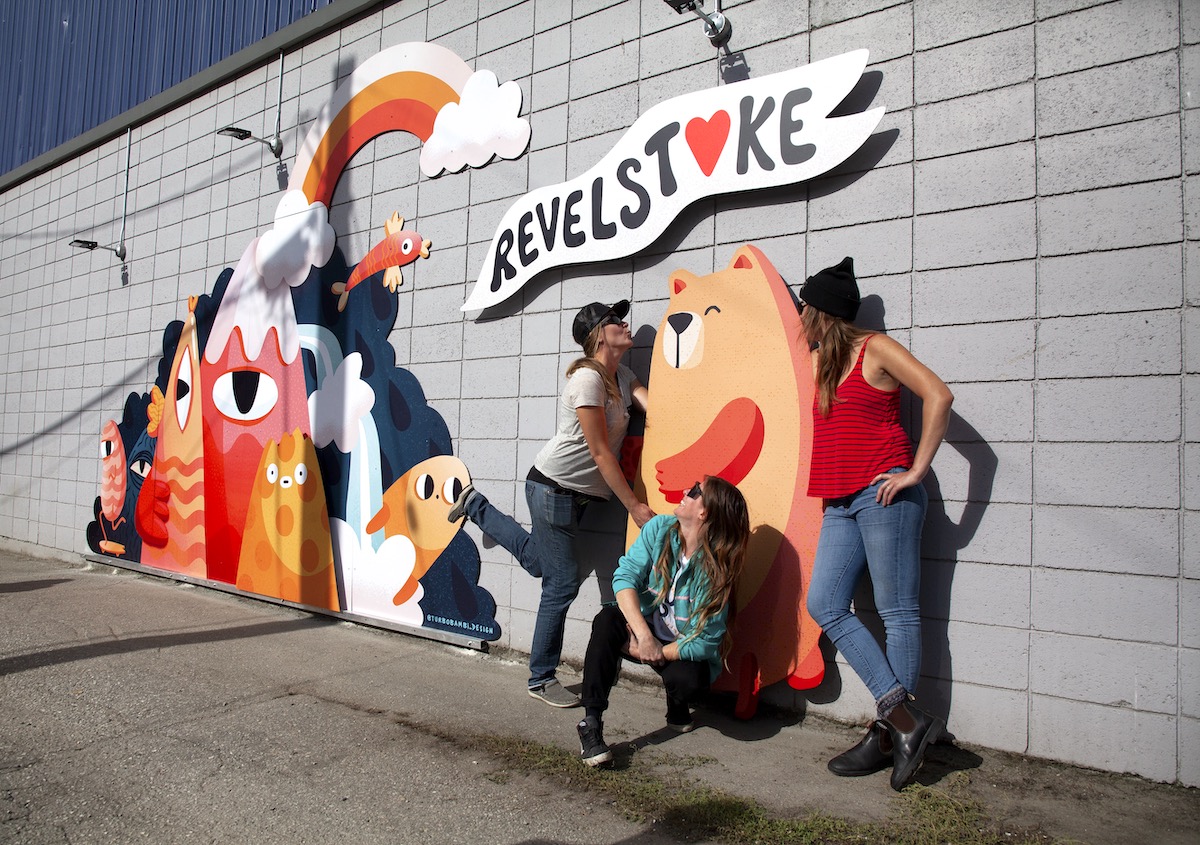 Three women stand in front of a mural with a bear under the word "Revelstoke". One pretends to kiss the bear.