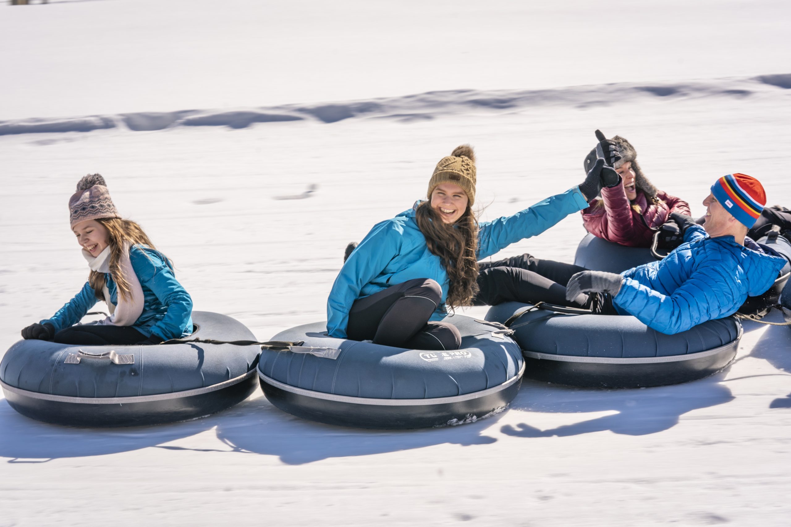 Four smiling people slide down a snowy hill on inner tubes.