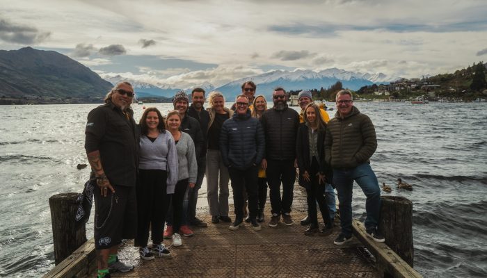 About a dozen people stand at the edge of a pier facgin the camera and smiling. Lake and mountains in the background.