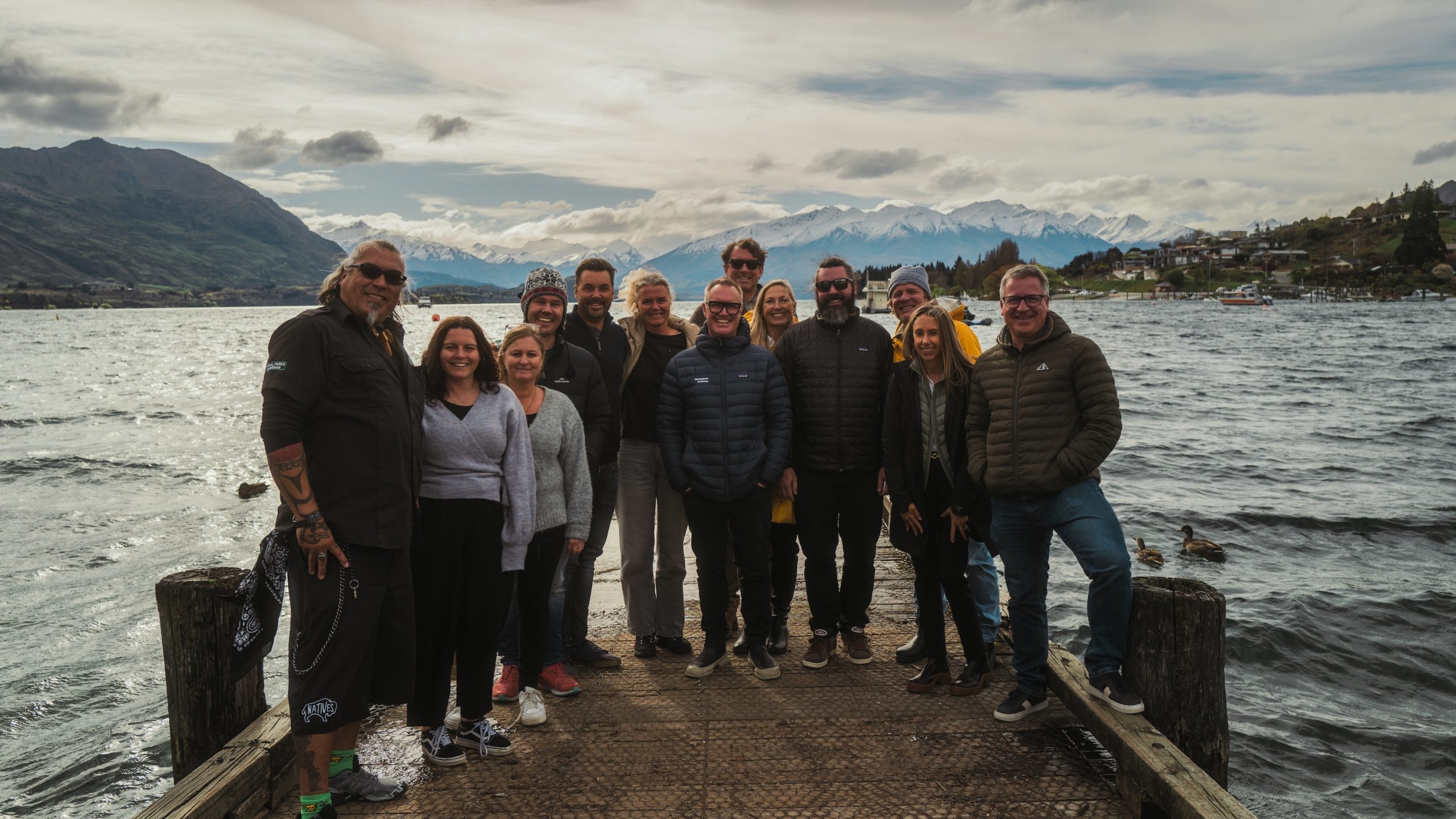 About a dozen people stand at the edge of a pier facgin the camera and smiling. Lake and mountains in the background.