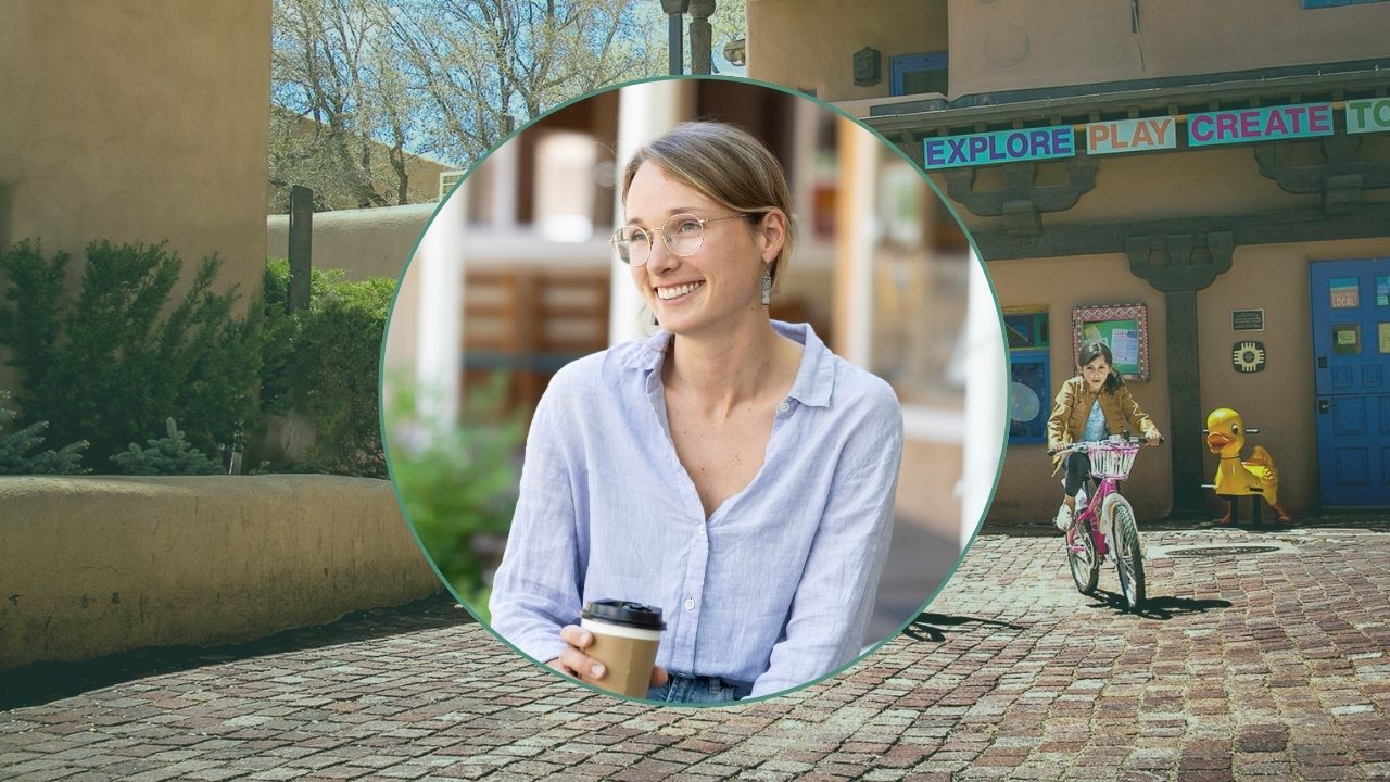 A white woman sits with a cup of coffee, smiling. Her photo is overlaid on a background of Taos, NM. Cobblestone streets and cyclists.