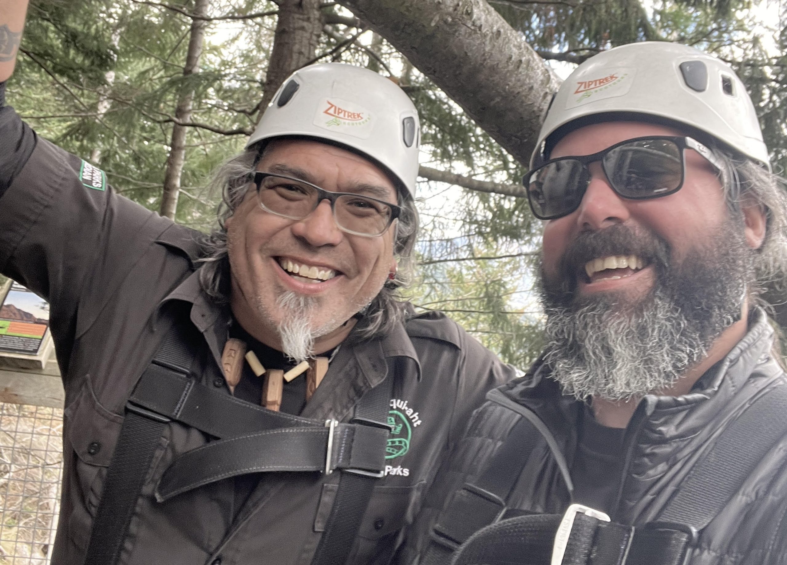 brad-saya-crop Two bearded men in white helmets and zipline gear in a forest smile at the camera.