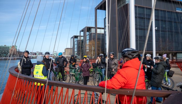 A bicycle tour group pauses to listen to the guide standing at a railing on the waterfront.