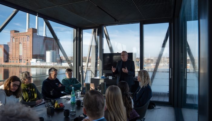 A man speaks to attendees in a meeting room with transparent walls. A factory is visible outside.
