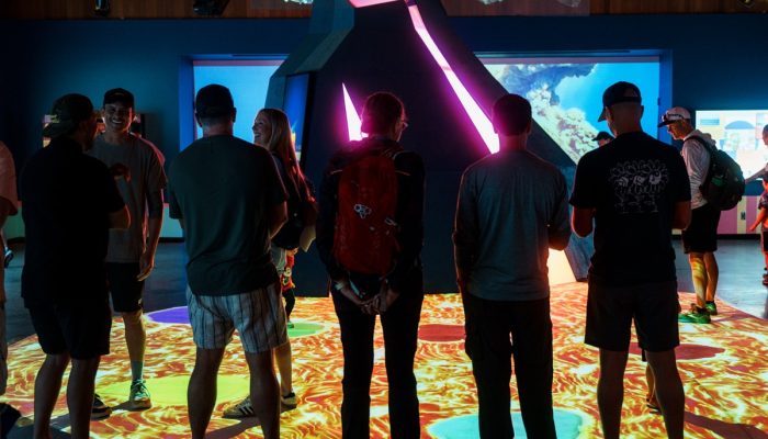 A group stands facing a volcano exhibit at a museum, backs to the camera.