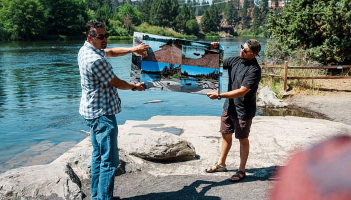 Two men hold images of the Deschutes River, standing next to the river.