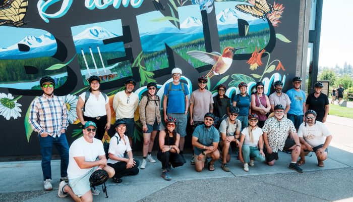 A group of about 20 people wearing bike helmets smiles in front of a "From BEND" mural.