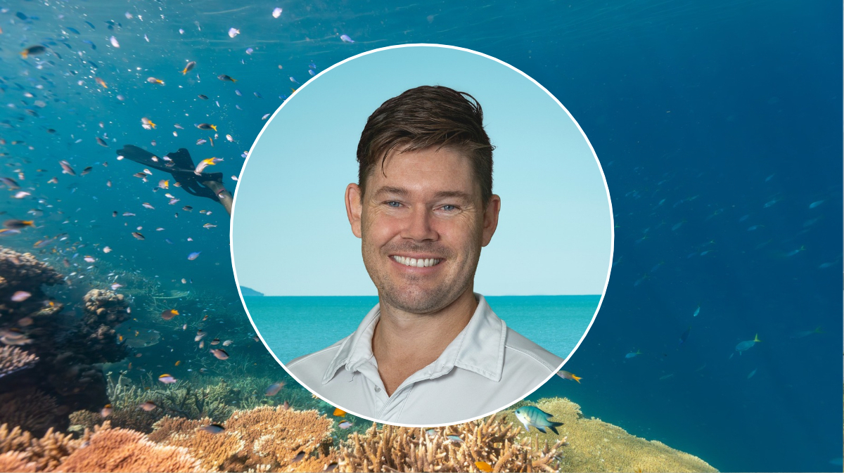 A smiling, light-skinned man looks toward the camera in a professional headshot. The background image shows colourful fish underwater.